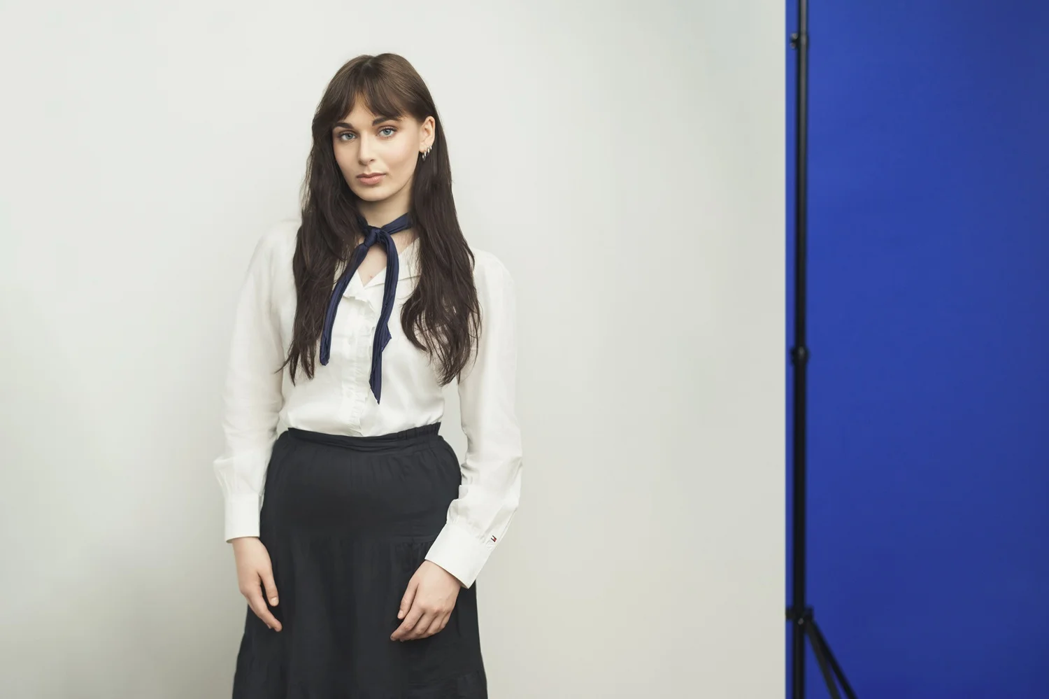 Young woman with long dark hair wearing a white shirt and navy neck scarf in a studio.