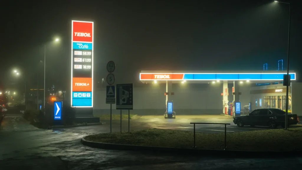 An illuminated gas station totem and canopy glow through thick night fog in a European setting.