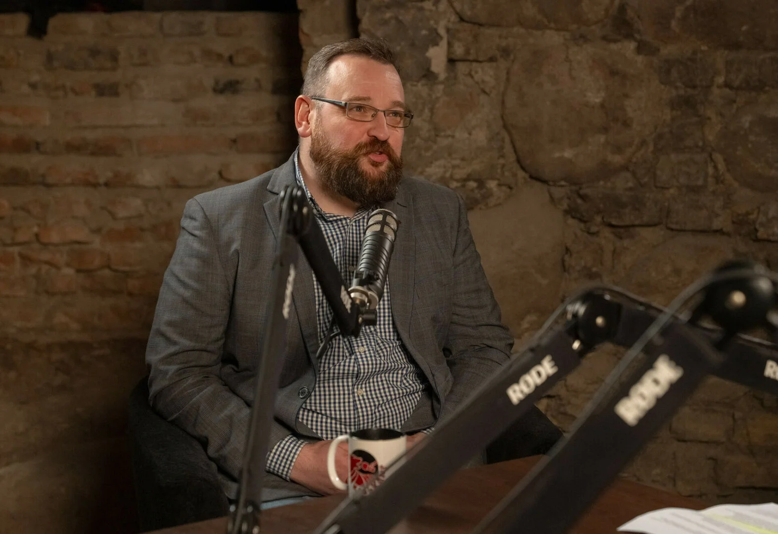 Bearded man in a blazer and glasses speaking into a professional microphone in a studio.