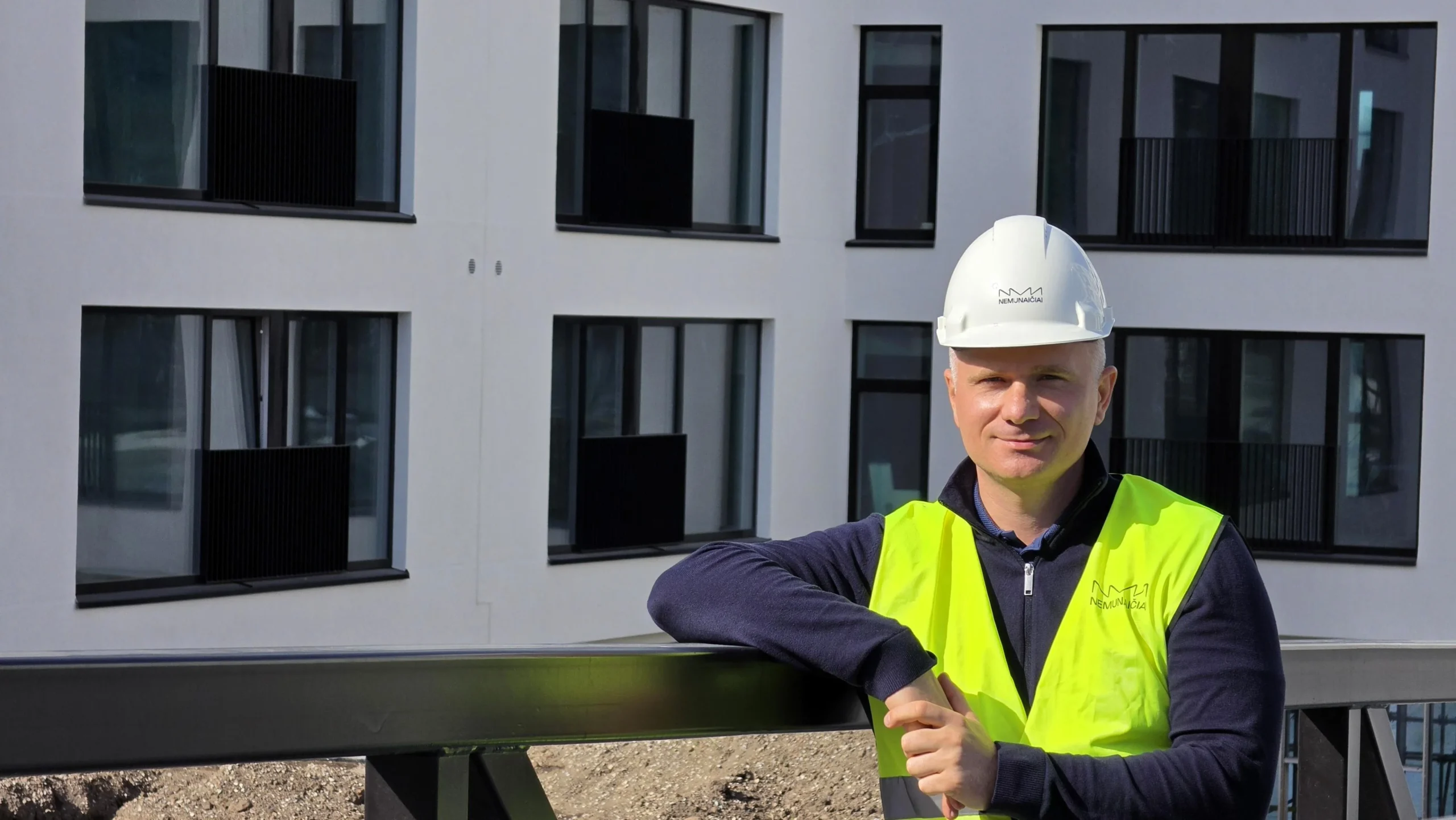 Man wearing white hard hat and yellow safety vest standing before a modern white apartment building.