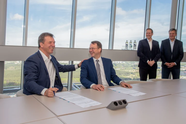 Two businessmen laughing while signing documents at a large conference table with a city view.