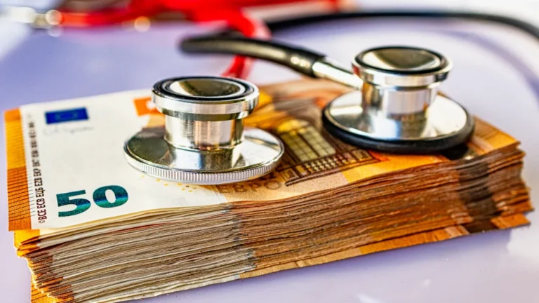 A medical stethoscope resting on a thick stack of fifty Euro banknotes on a bright surface.