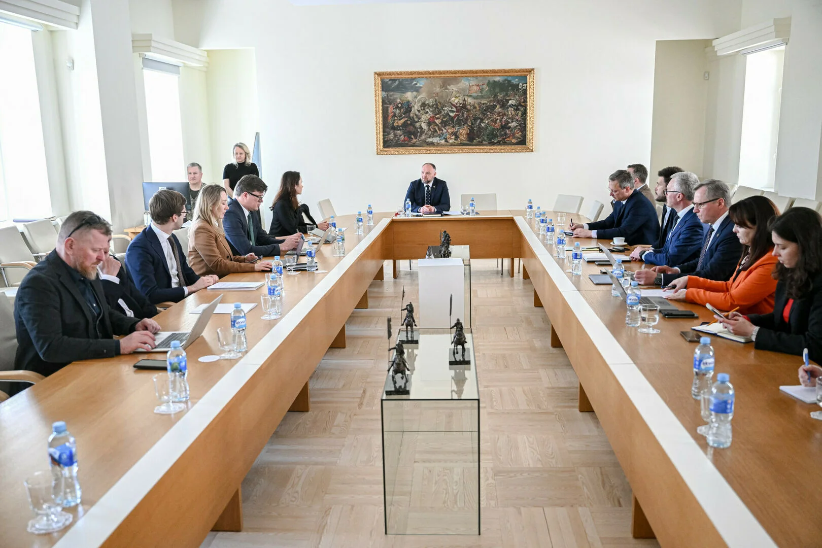 Officials in business suits sit around a large U-shaped wooden table during a formal conference meeting.