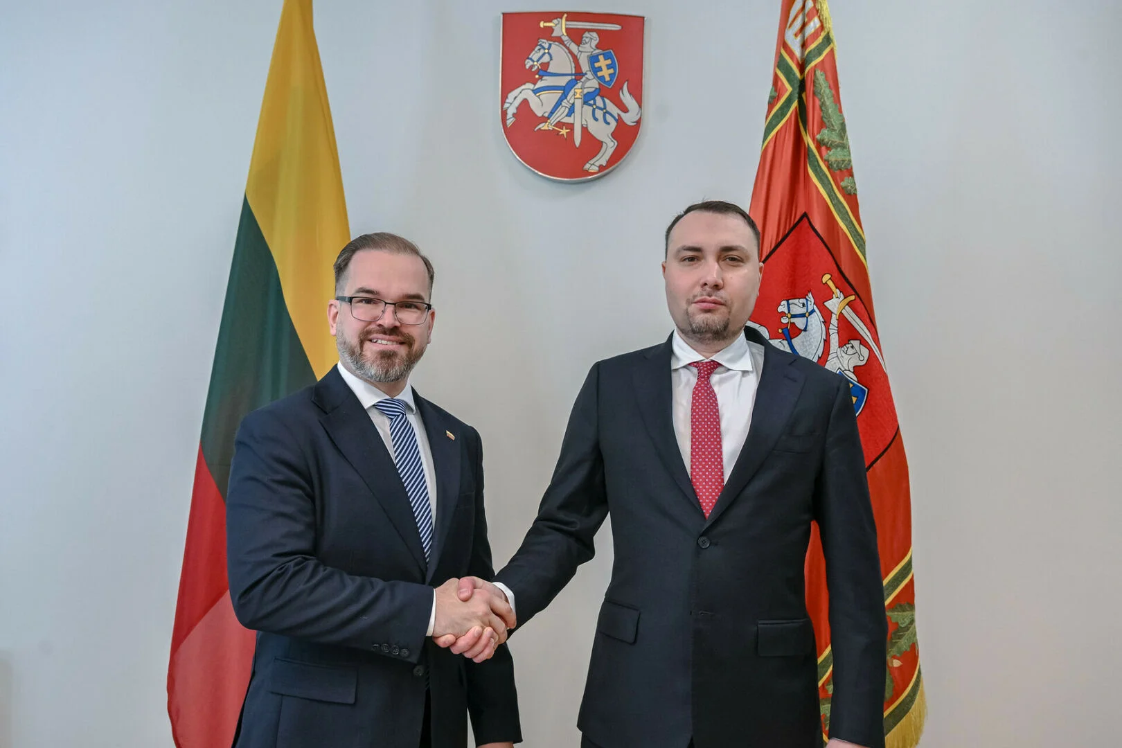 Two men in suits shaking hands before the Lithuanian flag and national coat of arms.