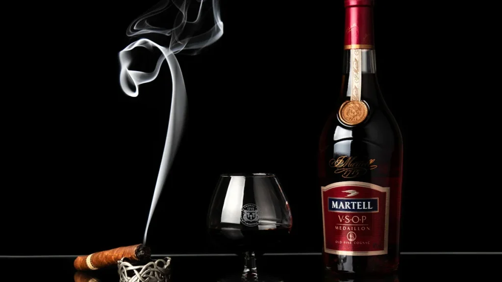 A bottle of cognac next to a glass and a lit cigar with rising smoke.