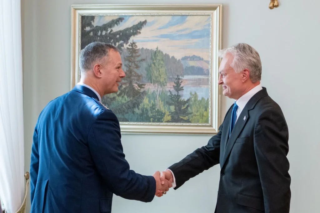 Military official in blue uniform shaking hands with a man in a suit before a landscape painting.