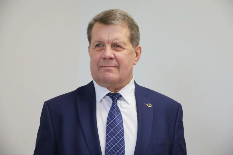 Middle-aged man wearing a navy suit and patterned blue tie against a plain light gray background.