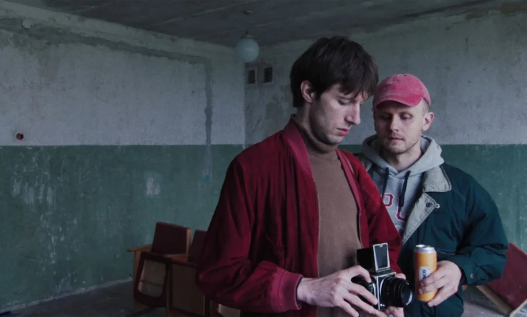 Two men in a dimly lit room, one adjusting a vintage camera and another holding a can.