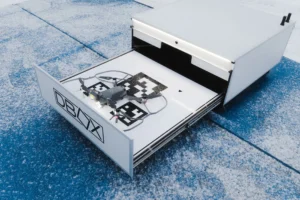 A gray drone sits on a patterned landing pad inside an open white DBOX storage drawer.