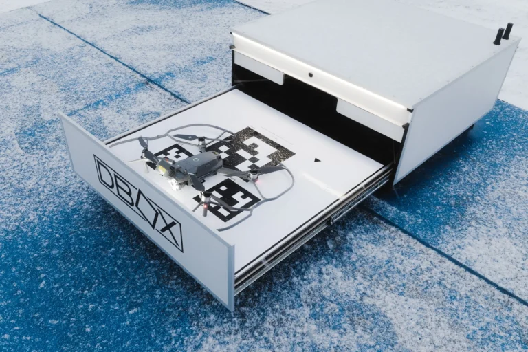 A gray drone sits on a patterned landing pad inside an open white DBOX storage drawer.