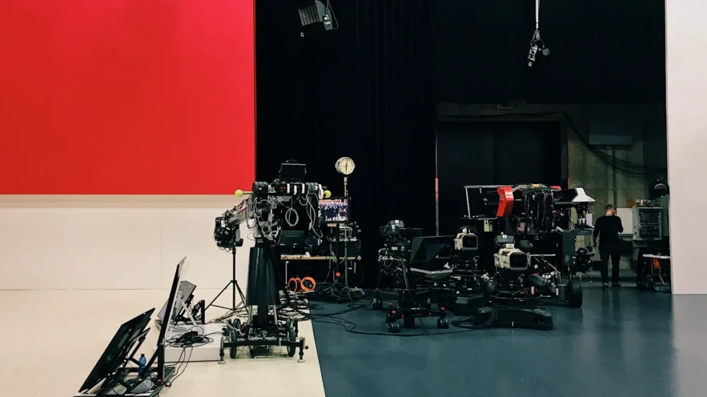 Professional television studio cameras and lighting equipment set up before a vibrant red and white backdrop.