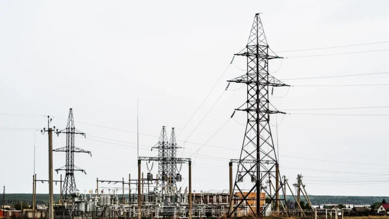 High-voltage electricity pylons and a transformer substation standing in a grassy rural field under overcast skies.