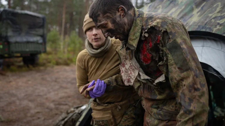 A military medic assists a simulated casualty with realistic moulage injuries during a tactical medicine training drill in a forest setting.