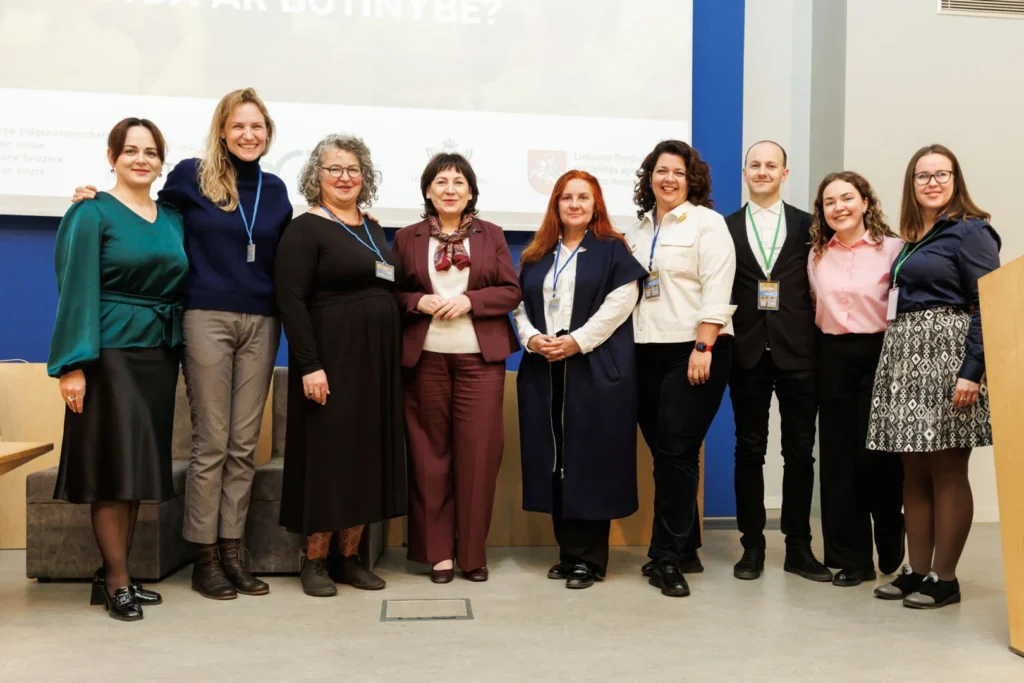 Nine smiling professionals stand together indoors in front of a white projection screen and blue wall.