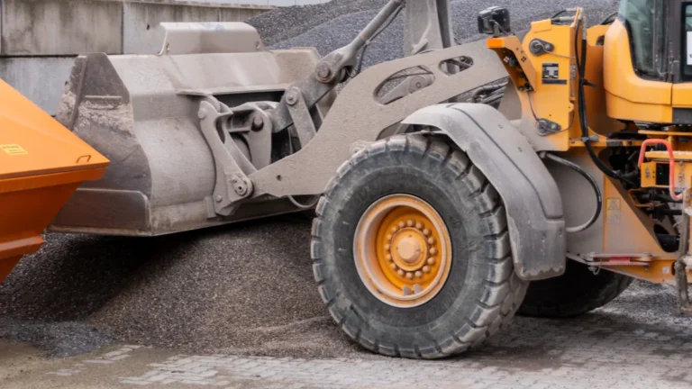 A yellow industrial loader scoops gravel into an orange truck at a European road construction site.