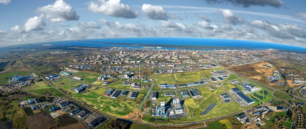 Wide aerial panorama of a coastal city featuring industrial zones, green fields, and the sea.