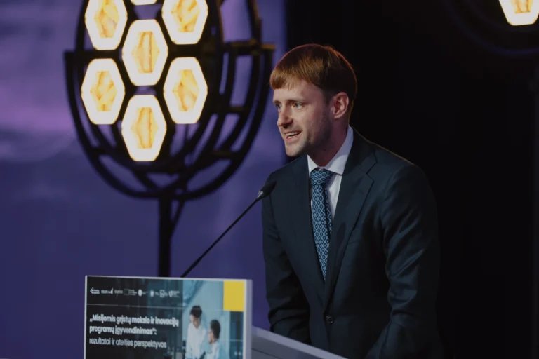 A man in a suit and patterned tie speaks at a podium with hexagonal lights.