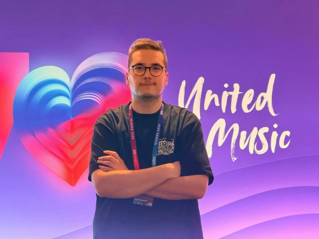 Man with glasses and crossed arms standing before a purple heart-themed 'United Music' backdrop.