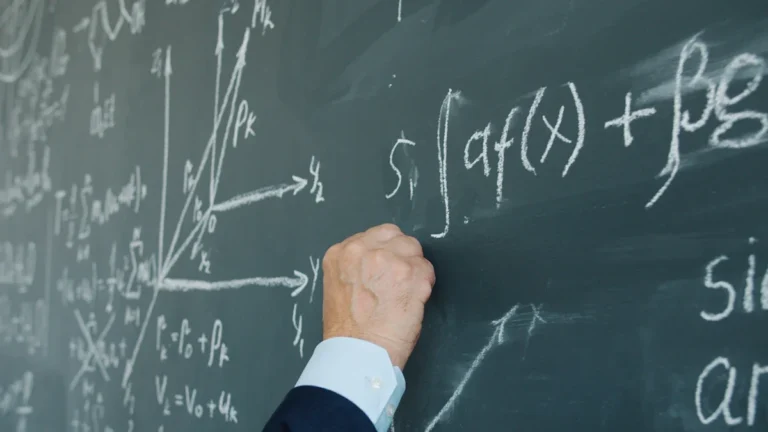 A close-up of a hand writing complex mathematical equations on a dark green chalkboard in a classroom.