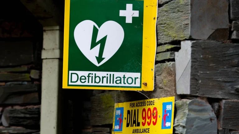 A green and yellow emergency defibrillator sign mounted on a stone wall with UK 999 instructions.