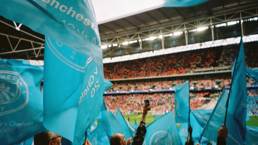 Manchester City fans waving light blue flags in a crowded football stadium.