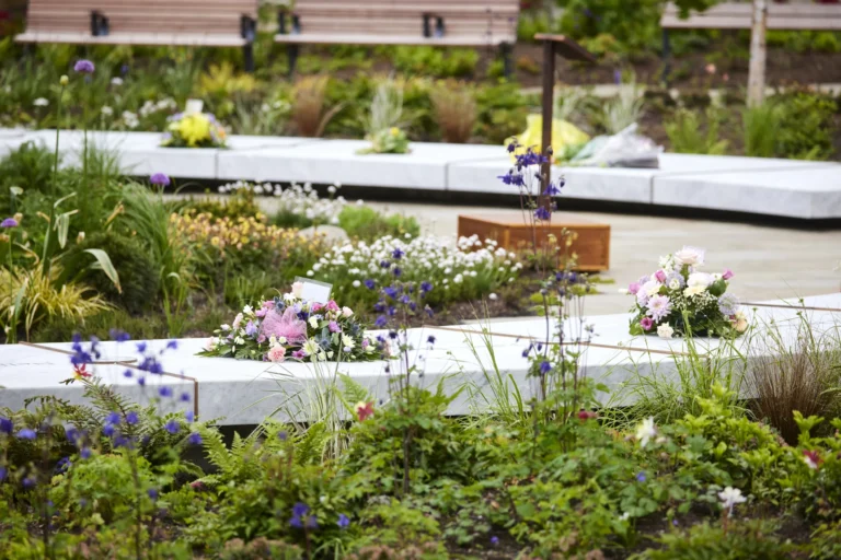 Floral tributes placed on white marble memorial benches surrounded by green plants and purple flowers.