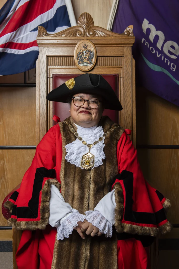A woman wearing red ceremonial mayoral robes and black hat sits in a wooden throne.