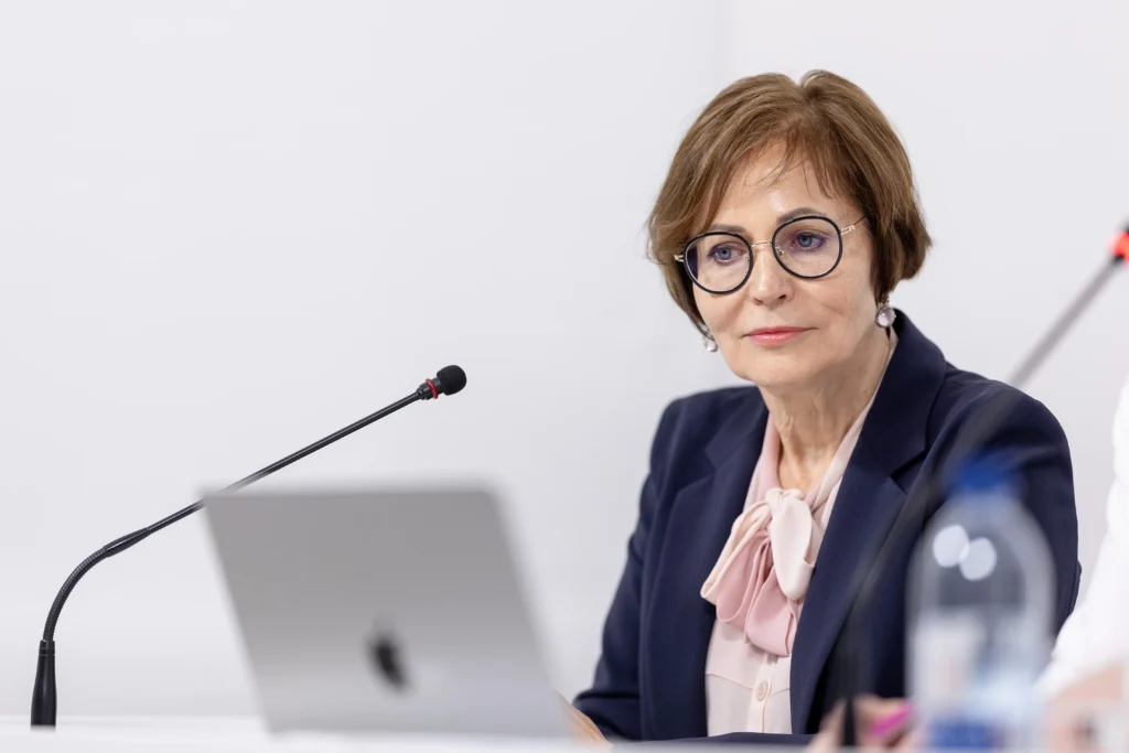 Professional woman with glasses wearing a navy blazer and pink bow blouse behind a microphone.