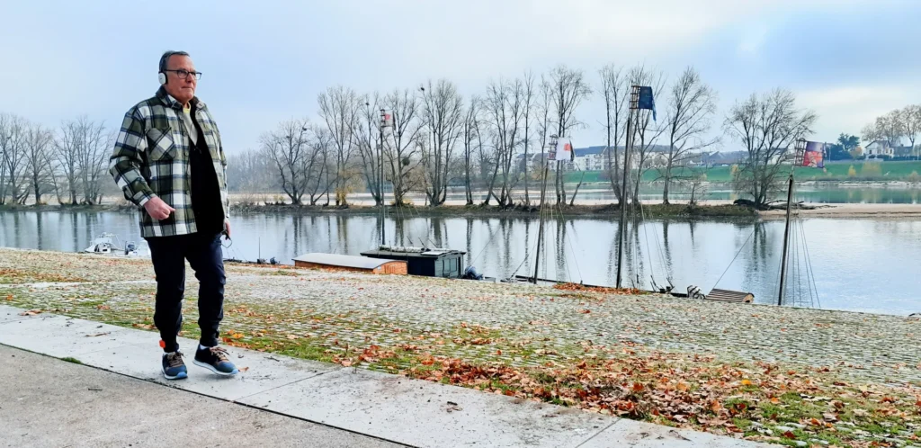 Older man in a plaid jacket walks along a riverbank with an electronic ankle monitor.