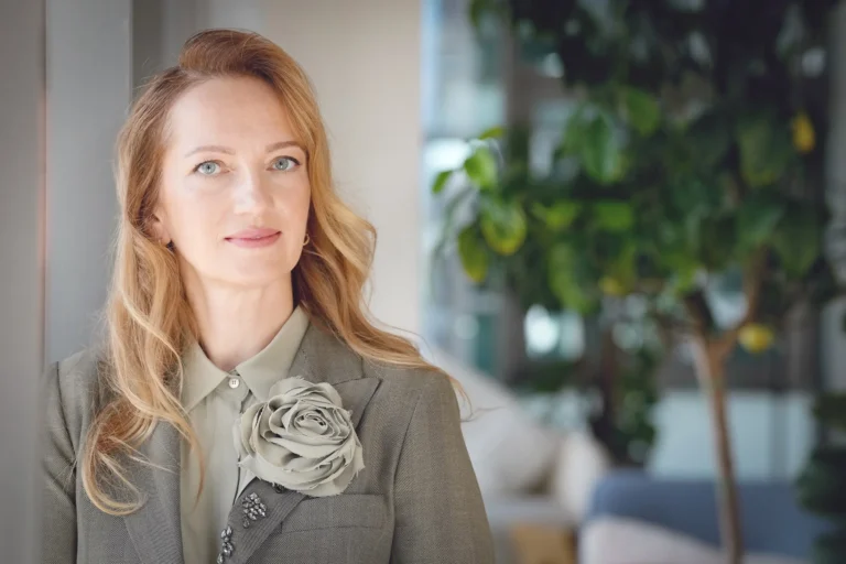 A blonde woman wearing a grey herringbone blazer and a large fabric flower brooch.