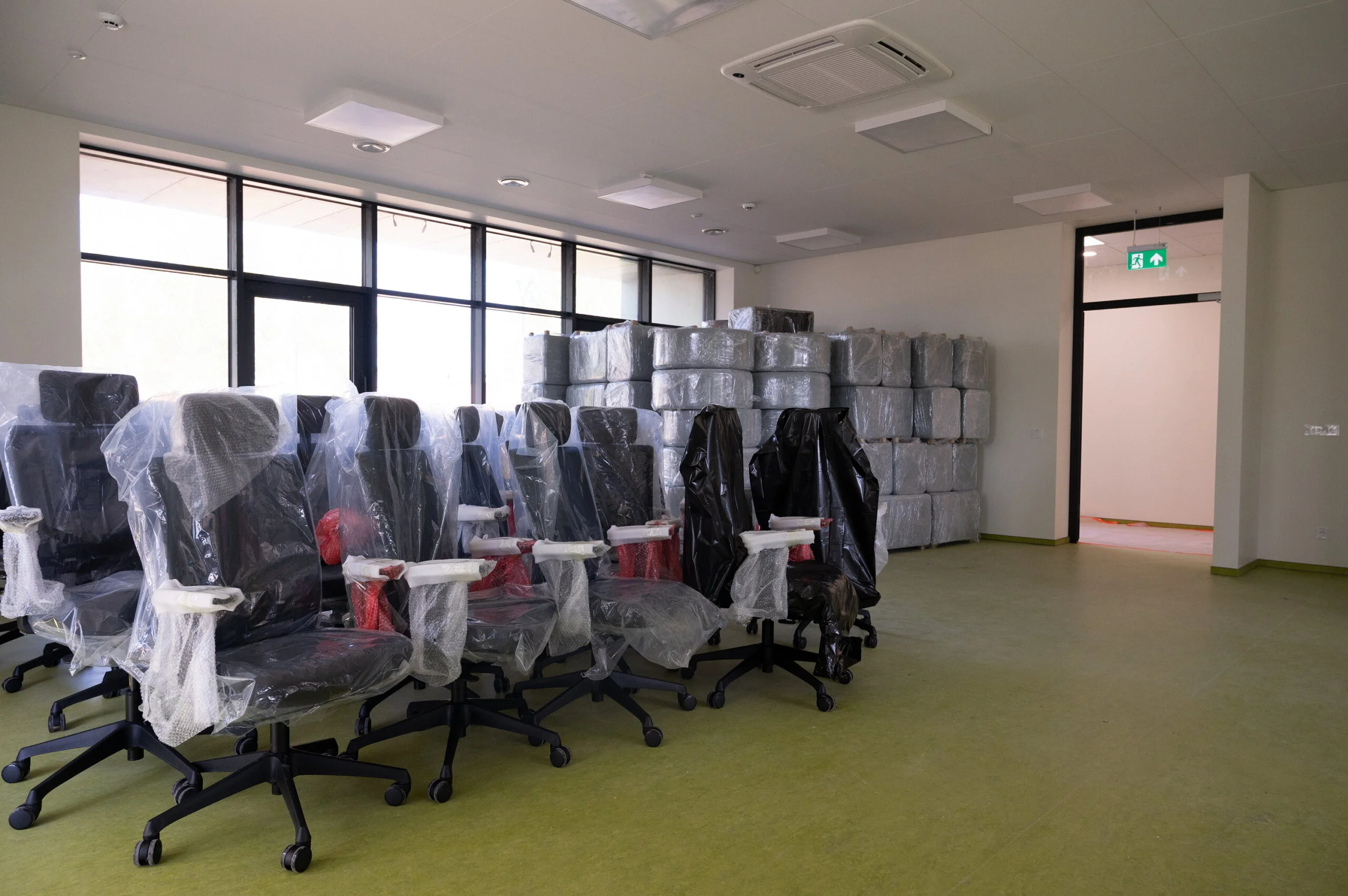 Rows of new black office chairs wrapped in protective plastic in a modern, sunlit workspace.