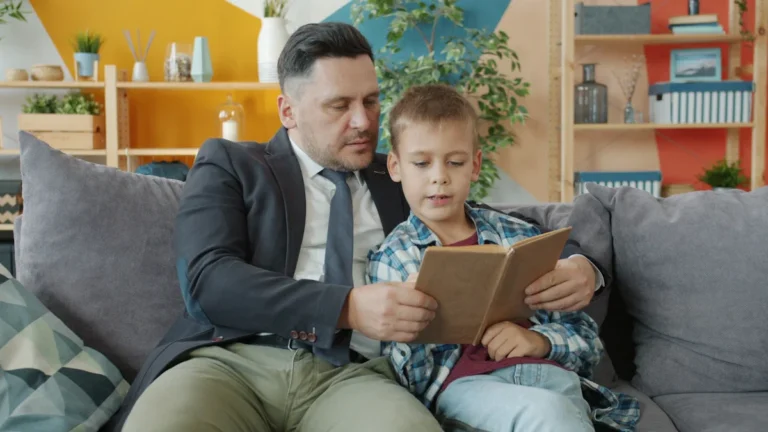 A man and a young boy sitting on a sofa and reading a book together in a brightly colored room.