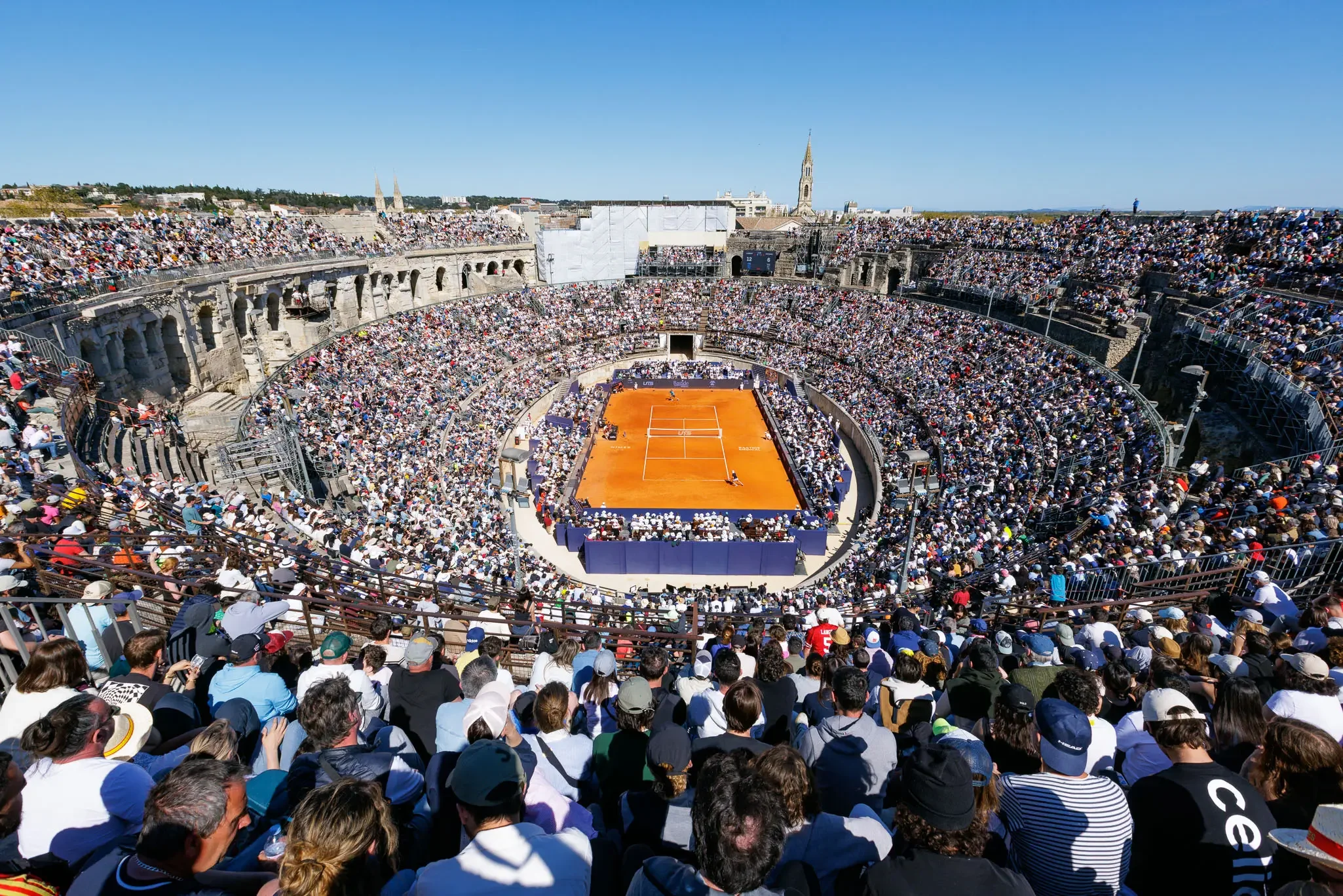 A clay tennis court inside a packed ancient Roman amphitheater under a clear blue sky.