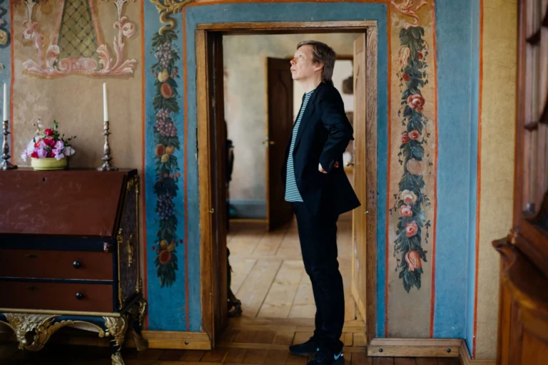 Man in a black blazer standing in an ornate doorway with painted floral wall decorations.