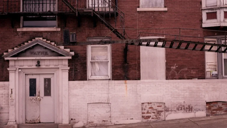 Exterior view of an old red brick apartment building with boarded-up windows and a white classical-style doorway.