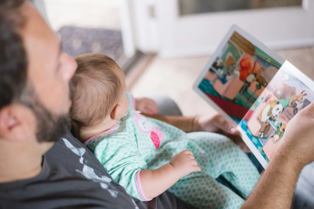 A man reading a colorful picture book to a baby sitting in his lap indoors.