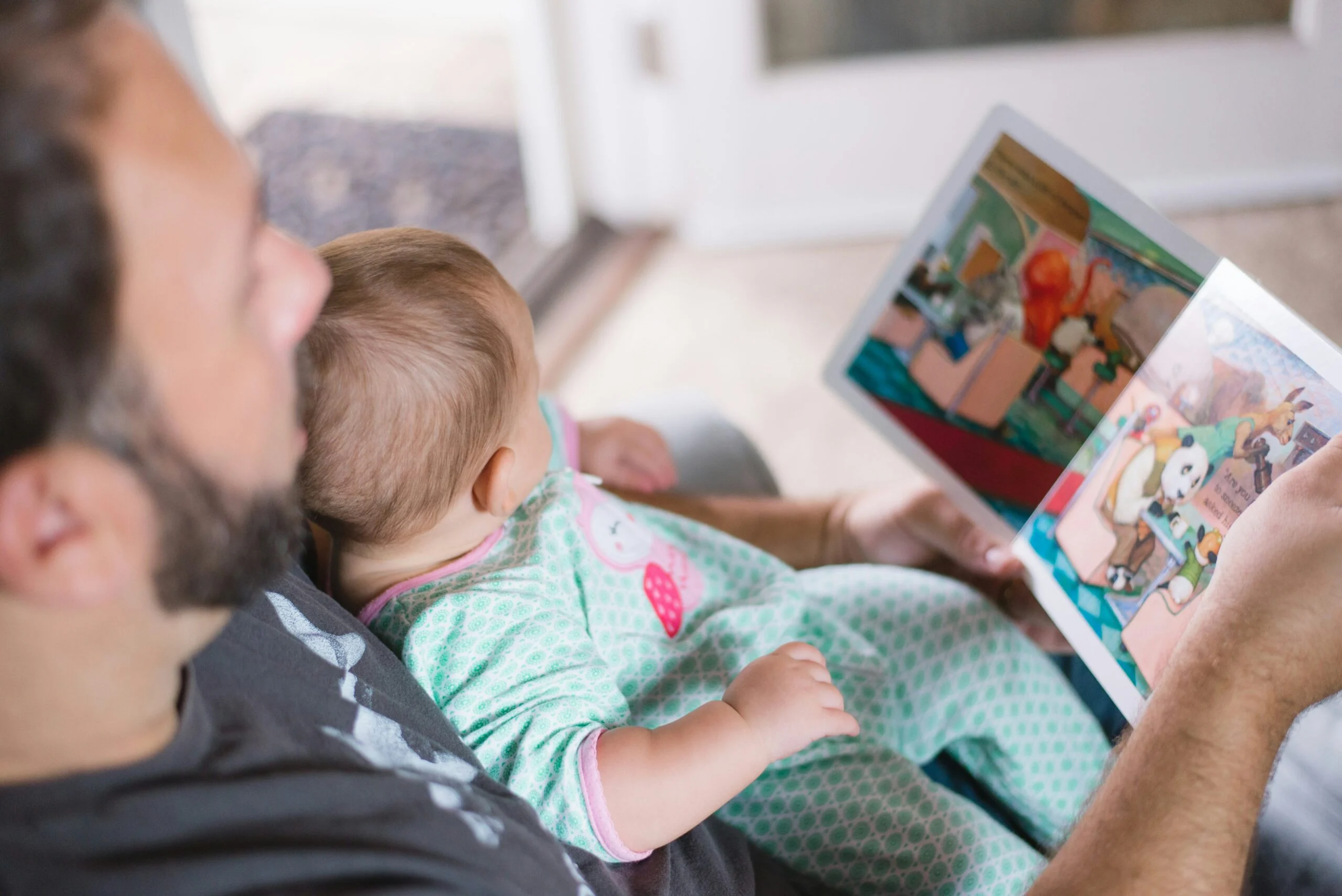 A man reading a colorful picture book to a baby sitting in his lap indoors.