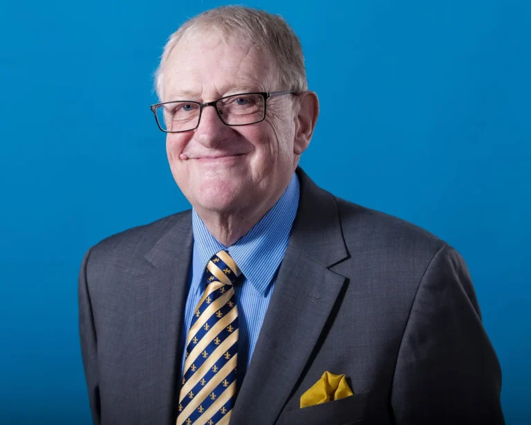 An older man with glasses wearing a grey suit and striped tie against blue.