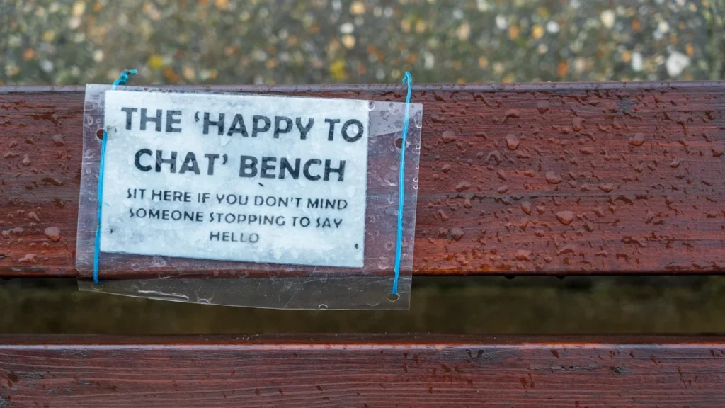 A close-up of a 'Happy to Chat' sign on a wet wooden park bench in the UK.
