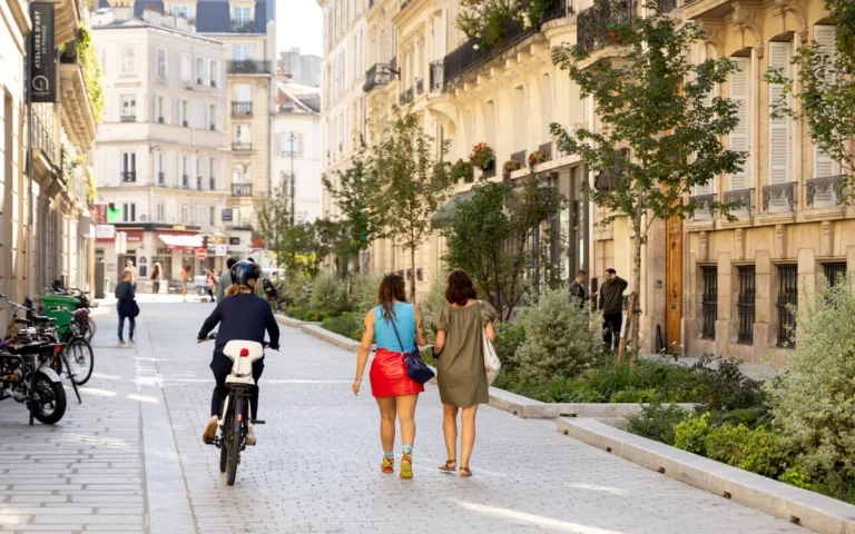 People walk and cycle down a sunny, paved street lined with trees and historic city architecture.