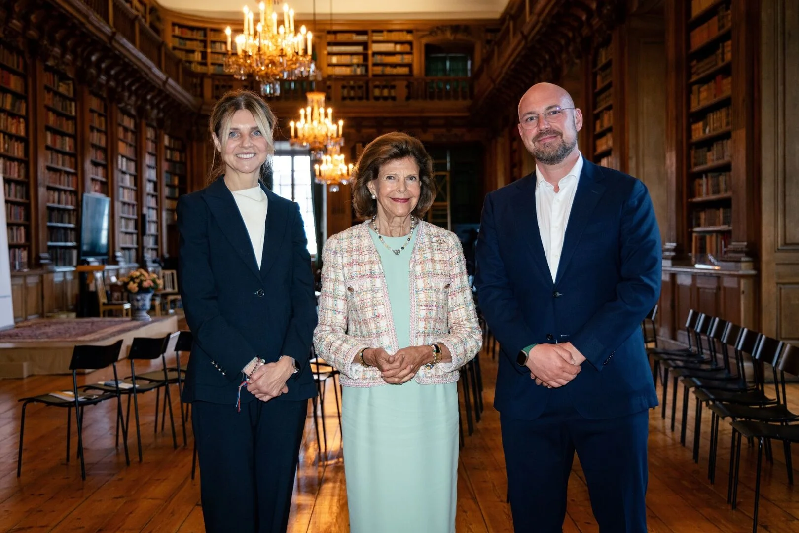Queen Silvia of Sweden stands with two professionals inside a grand, historic library filled with books.