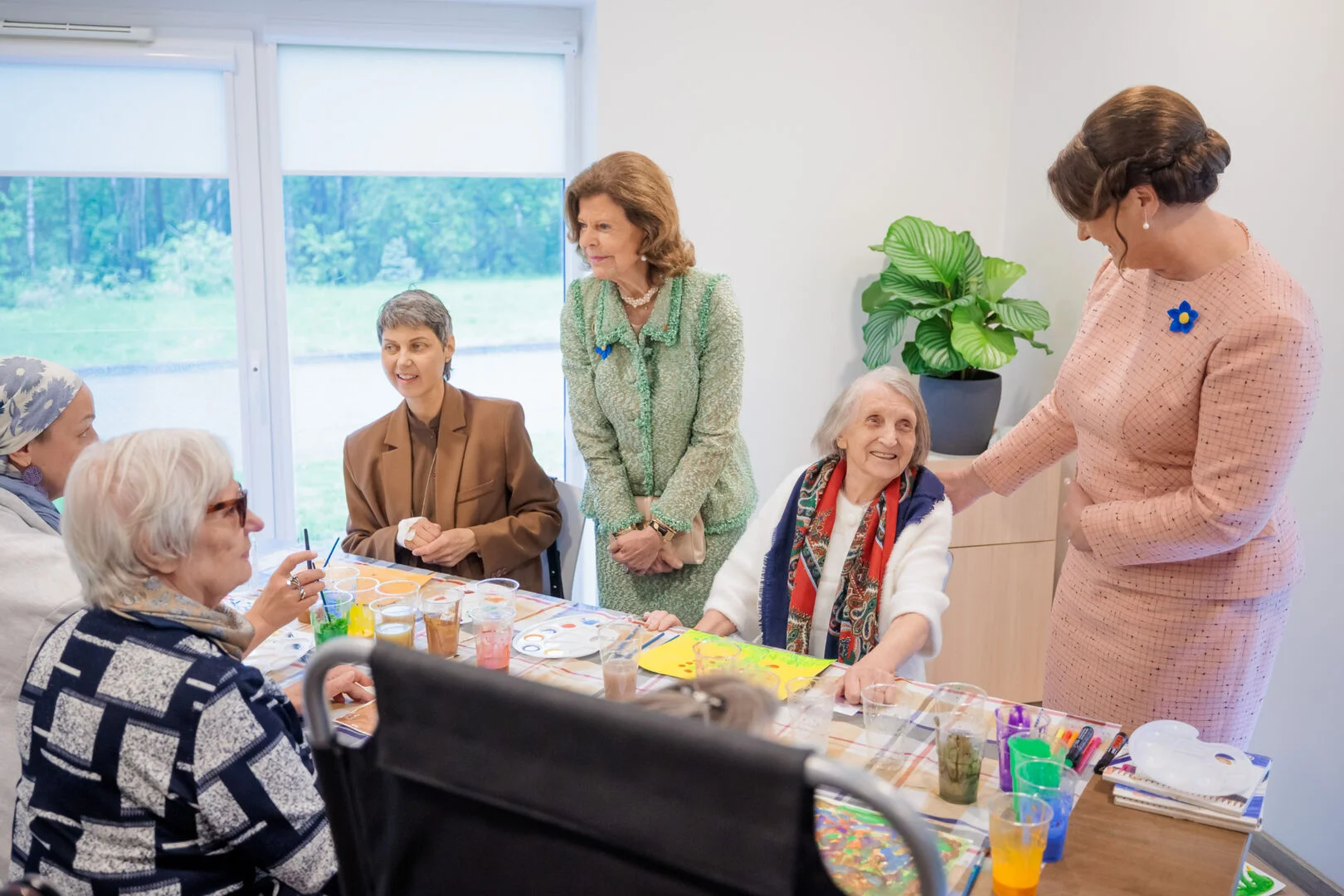 Dignitaries in formal suits visit seniors painting at a table in a brightly lit room.