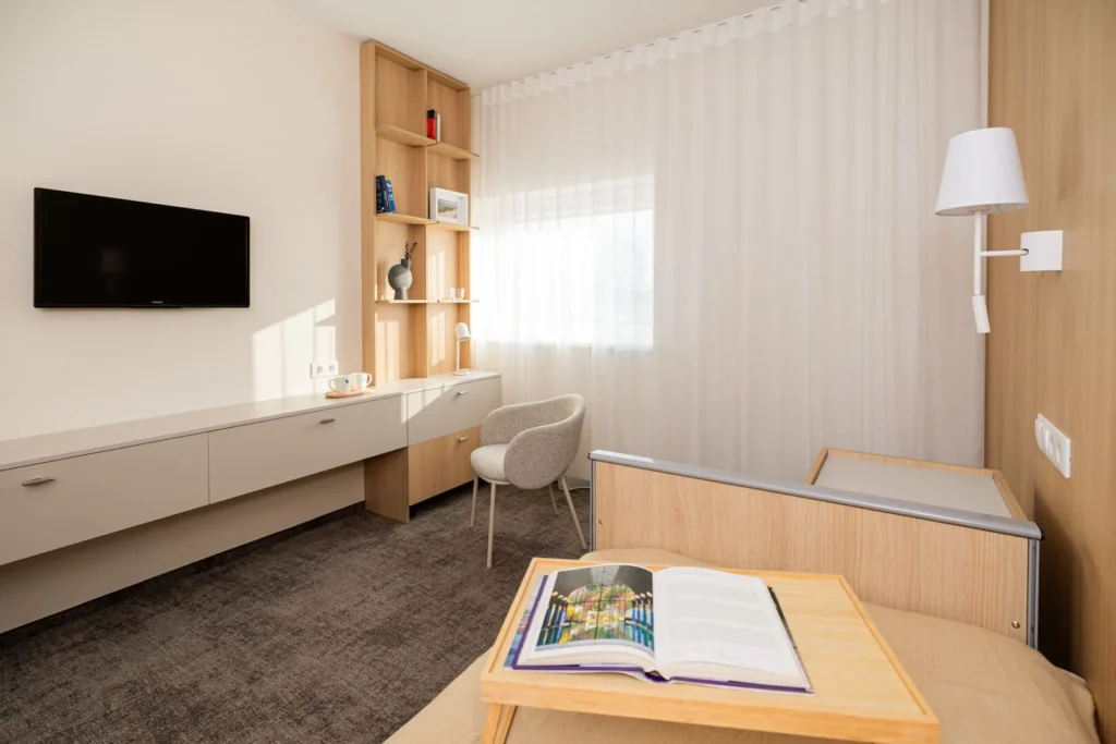 Modern minimalist care room featuring light wood furniture, a wall-mounted TV, and an armchair.