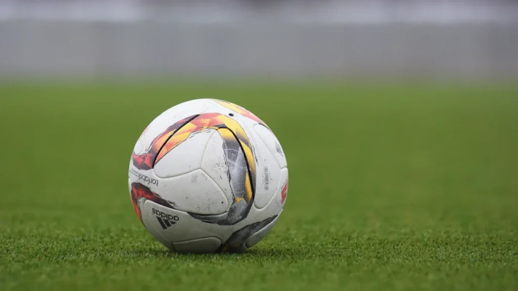 An Adidas soccer ball sits on a green grass pitch during the day.