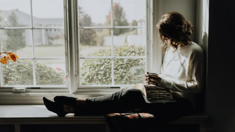A woman sitting peacefully on a window sill with a book and coffee, reflecting on her healing journey.