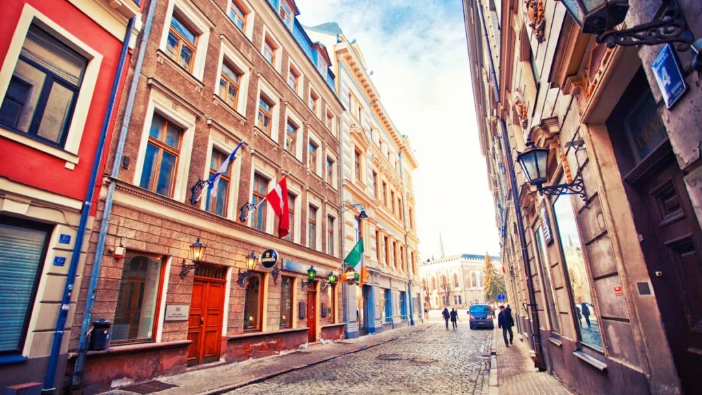 A historic cobblestone street in Latvia featuring colorful classic architecture and the national flag.