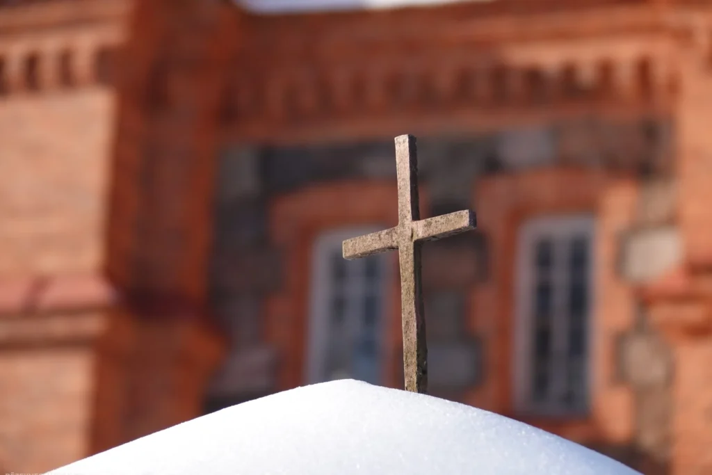 Small stone cross standing in fresh white snow with a blurred red brick building background.