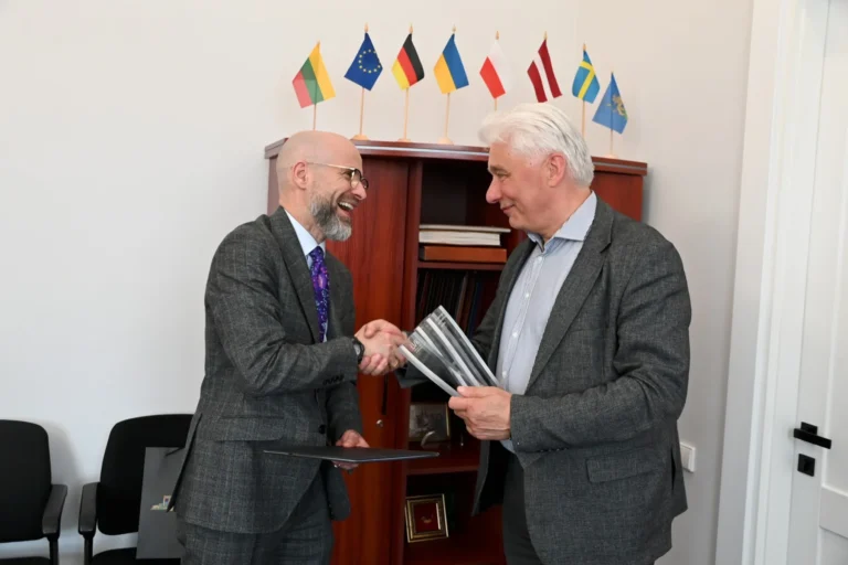Two men in grey suits shaking hands and laughing in front of various national flags.