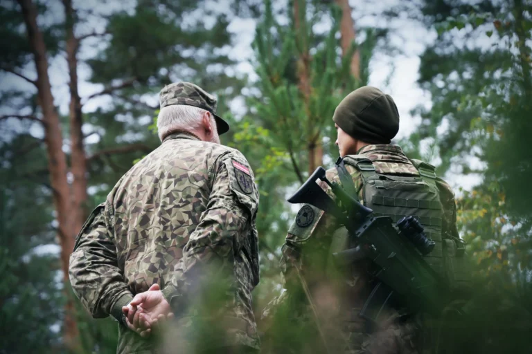 Older man and younger soldier in camouflage uniforms stand together in a wooded forest area.