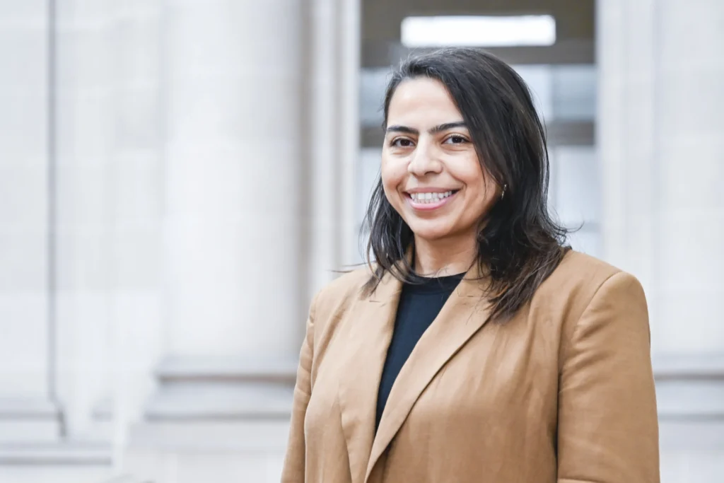A smiling woman with dark hair wearing a tan blazer in front of a building.